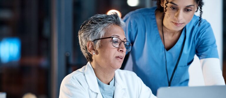 Deux médecins femme de face regardant un ordinateur (l'une assise, l'autre penchée au dessus de son épaule)
