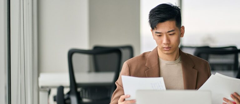 Jeune homme asiatique de face assis à droite d'une table devant un ordinateur et regardant des documents dans ses mains